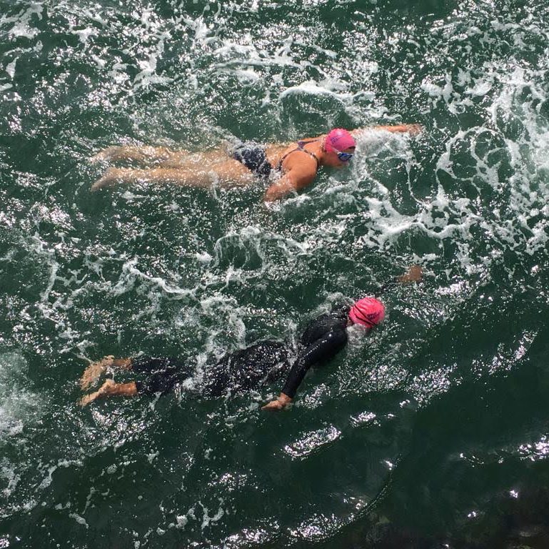 Photo of two swimmers in the sea. Both are wearing pink swim hats and goggles, one is wearing a wetsuit, and the other is wearing a swimsuit.